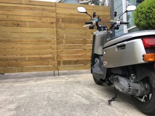 A modern scooter is parked on a concrete driveway in front of a wooden fence. The scooter is metallic silver with black accents, and its rear wheel and exhaust system are visible. In the background, there is a bush next to a window, partially reflecting the surroundings.