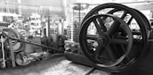 A collection of industrial machinery and equipment is scattered in a workshop setting. Prominently featured is a large flywheel with several smaller gears and mechanical components around it. There are gas cylinders in the background and various metal and wooden planks are visible, creating a cluttered but industrious atmosphere.