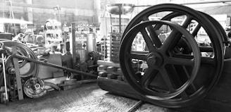 A collection of industrial machinery and equipment is scattered in a workshop setting. Prominently featured is a large flywheel with several smaller gears and mechanical components around it. There are gas cylinders in the background and various metal and wooden planks are visible, creating a cluttered but industrious atmosphere.