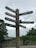 A wooden signpost with multiple directional signs pointing towards various village attractions such as the Village Hall, Post Office, Car Park, Shop and Information, Grove Rare Books, Lavatories, and The Tea Cottage. The background features greenery and a stone wall under a cloudy sky.