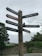 A wooden signpost with multiple directional signs pointing towards various village attractions such as the Village Hall, Post Office, Car Park, Shop and Information, Grove Rare Books, Lavatories, and The Tea Cottage. The background features greenery and a stone wall under a cloudy sky.