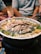 A hotpot filled with a variety of ingredients including sliced meat, green vegetables, white balls possibly fish or meat balls, and other assorted items. The hotpot is set on a table with a spoon nearby and some people in the background.