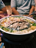A hotpot filled with a variety of ingredients including sliced meat, green vegetables, white balls possibly fish or meat balls, and other assorted items. The hotpot is set on a table with a spoon nearby and some people in the background.