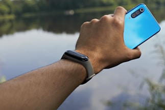 A hand with a fitness watch is capturing a photo using a blue smartphone near a calm lake, with a blurred background of trees and water.