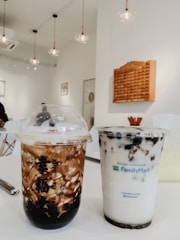 Two bubble tea drinks are placed on a white table in a modern cafe setting. The drink on the left has a darker, swirling pattern with black tapioca pearls at the bottom, and the drink on the right has a light, creamy appearance with black tapioca pearls. The cafe interior features minimalist decor with hanging light fixtures, white walls, and framed art.