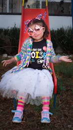A young child sits on a red slide wearing colorful and whimsical clothing. The outfit includes a rainbow cardigan, a black T-shirt with playful text, a white tutu, and striped leg warmers. The child accessorizes with oversized pink sunglasses adorned with sparkles and a headband featuring shiny star decorations. Blue and pink sneakers complete the vibrant look.