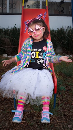 A young child sits on a red slide wearing colorful and whimsical clothing. The outfit includes a rainbow cardigan, a black T-shirt with playful text, a white tutu, and striped leg warmers. The child accessorizes with oversized pink sunglasses adorned with sparkles and a headband featuring shiny star decorations. Blue and pink sneakers complete the vibrant look.