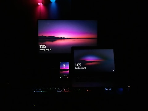 A dark room featuring multiple screens, including a television, a laptop, and a smartphone, all displaying the same vivid wallpaper of a serene landscape at dusk. The wallpaper features a prominent gradient of pink, purple, and blue colors. Soft ambient lighting from red, blue, and green LED lights adds to the atmosphere.