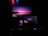 A dark room featuring multiple screens, including a television, a laptop, and a smartphone, all displaying the same vivid wallpaper of a serene landscape at dusk. The wallpaper features a prominent gradient of pink, purple, and blue colors. Soft ambient lighting from red, blue, and green LED lights adds to the atmosphere.
