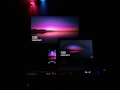 A dark room featuring multiple screens, including a television, a laptop, and a smartphone, all displaying the same vivid wallpaper of a serene landscape at dusk. The wallpaper features a prominent gradient of pink, purple, and blue colors. Soft ambient lighting from red, blue, and green LED lights adds to the atmosphere.