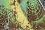 A rusted metal surface is covered with peeling paint in shades of green and yellow. The layers of deteriorating paint create a textured pattern, while a spiral design in bright green adds an artistic element to the weathered surface.
