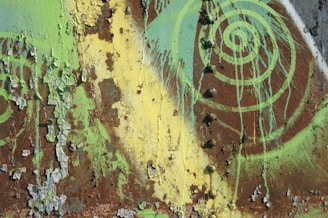 A rusted metal surface is covered with peeling paint in shades of green and yellow. The layers of deteriorating paint create a textured pattern, while a spiral design in bright green adds an artistic element to the weathered surface.