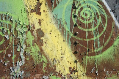 A rusted metal surface is covered with peeling paint in shades of green and yellow. The layers of deteriorating paint create a textured pattern, while a spiral design in bright green adds an artistic element to the weathered surface.