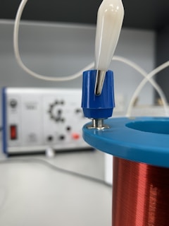 A close-up view of an electrical coil wrapped with copper wire, placed in the foreground. The coil has a blue and red casing, with a white and blue connector attached to the top. In the blurred background, there is electronic equipment, possibly a control panel or an oscilloscope, featuring various knobs and connectors.