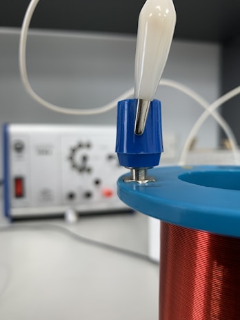 A close-up view of an electrical coil wrapped with copper wire, placed in the foreground. The coil has a blue and red casing, with a white and blue connector attached to the top. In the blurred background, there is electronic equipment, possibly a control panel or an oscilloscope, featuring various knobs and connectors.