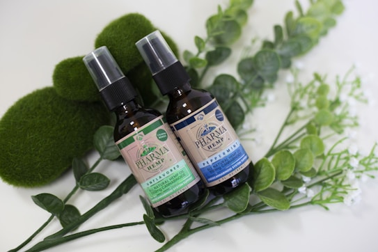 Two brown spray bottles labeled 'Pharma Hemp' placed on a bed of green leaves and surrounded by mossy and leafy decorations. One bottle is marked 'unflavored' and the other 'peppermint'. The layout is aesthetically arranged with a natural, organic vibe.