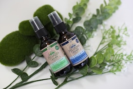 Two brown spray bottles labeled 'Pharma Hemp' placed on a bed of green leaves and surrounded by mossy and leafy decorations. One bottle is marked 'unflavored' and the other 'peppermint'. The layout is aesthetically arranged with a natural, organic vibe.