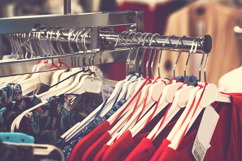 A clothing rack filled with various garments on hangers, with a prominent display of red and floral patterns. The focus is on neatly organized clothes in a retail setting, highlighting the variety and arrangement.