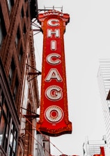 A vertical red sign attached to a building displays the word 'CHICAGO' in large, illuminated letters. The sign is ornate with intricate details and is positioned against a backdrop of urban architecture, including modern skyscrapers and an older brick building with decorative elements.