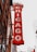 A vertical red sign attached to a building displays the word 'CHICAGO' in large, illuminated letters. The sign is ornate with intricate details and is positioned against a backdrop of urban architecture, including modern skyscrapers and an older brick building with decorative elements.