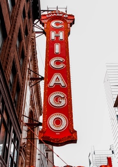A vertical red sign attached to a building displays the word 'CHICAGO' in large, illuminated letters. The sign is ornate with intricate details and is positioned against a backdrop of urban architecture, including modern skyscrapers and an older brick building with decorative elements.