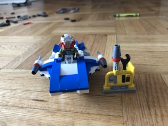 A small Lego spacecraft with a mini-figure wearing a helmet sits on a wooden floor. The spacecraft is predominantly blue and white with two grey and red thrusters. Next to it, there is a small structure with tools, including a wrench, standing on a grey and yellow base. Scattered Lego pieces and a crayon are visible in the background.