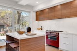 A modern kitchen features wooden cabinetry and a vibrant red oven range. The countertops are sleek white marble, complemented by a matching backsplash. An island with a wood base and white countertop includes two wooden stools. Decorative elements like a dark vase and a wooden bowl add a touch of elegance. Large windows allow natural light in, revealing a view of a backyard with trees and a grill.