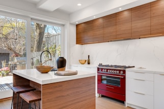 A modern kitchen features wooden cabinetry and a vibrant red oven range. The countertops are sleek white marble, complemented by a matching backsplash. An island with a wood base and white countertop includes two wooden stools. Decorative elements like a dark vase and a wooden bowl add a touch of elegance. Large windows allow natural light in, revealing a view of a backyard with trees and a grill.