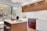 A modern kitchen features wooden cabinetry and a vibrant red oven range. The countertops are sleek white marble, complemented by a matching backsplash. An island with a wood base and white countertop includes two wooden stools. Decorative elements like a dark vase and a wooden bowl add a touch of elegance. Large windows allow natural light in, revealing a view of a backyard with trees and a grill.