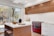 A modern kitchen features wooden cabinetry and a vibrant red oven range. The countertops are sleek white marble, complemented by a matching backsplash. An island with a wood base and white countertop includes two wooden stools. Decorative elements like a dark vase and a wooden bowl add a touch of elegance. Large windows allow natural light in, revealing a view of a backyard with trees and a grill.