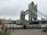 Tower Bridge in London with its distinctive twin towers and blue suspension components. The overcast sky creates a muted backdrop. People are walking near the riverbank, and tall grasses are seen in the foreground.