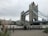Tower Bridge in London with its distinctive twin towers and blue suspension components. The overcast sky creates a muted backdrop. People are walking near the riverbank, and tall grasses are seen in the foreground.