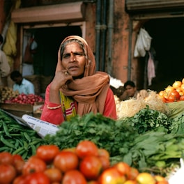 A woman in a traditional headscarf sits at a market stall surrounded by a variety of fresh vegetables including tomatoes, green beans, and leafy greens. She appears thoughtful with her hand resting on her chin. Other people are visible in the background, and the setting is a busy market street with a rustic ambiance.