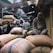 A bustling marketplace with several people engaged in various activities. A man sits atop large burlap sacks filled with goods, wrapped in a shawl, observing the surroundings. The market has a narrow pathway with crowded stalls and a canopy of makeshift coverings. The atmosphere appears lively with a focus on trade and commerce.