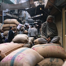 A bustling marketplace with several people engaged in various activities. A man sits atop large burlap sacks filled with goods, wrapped in a shawl, observing the surroundings. The market has a narrow pathway with crowded stalls and a canopy of makeshift coverings. The atmosphere appears lively with a focus on trade and commerce.