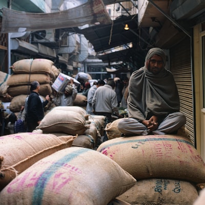 A bustling marketplace with several people engaged in various activities. A man sits atop large burlap sacks filled with goods, wrapped in a shawl, observing the surroundings. The market has a narrow pathway with crowded stalls and a canopy of makeshift coverings. The atmosphere appears lively with a focus on trade and commerce.
