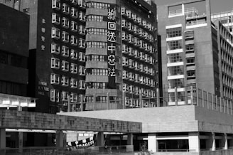 A black and white image of urban buildings with many windows and visible signage. The architecture features a mix of modern and older styles, and some text on the buildings is visible. The scene captures a complex cityscape.
