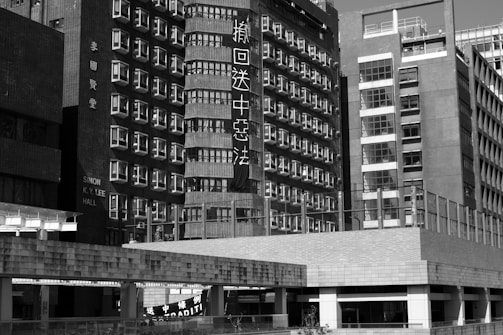 A black and white image of urban buildings with many windows and visible signage. The architecture features a mix of modern and older styles, and some text on the buildings is visible. The scene captures a complex cityscape.