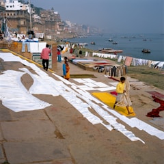 A riverside scene with people drying laundry on the ghats. White sheets are spread out on the ground, and colorful clothes hang on lines. The river has several boats floating, and the buildings line the bank, showing a dense and historic architectural style.