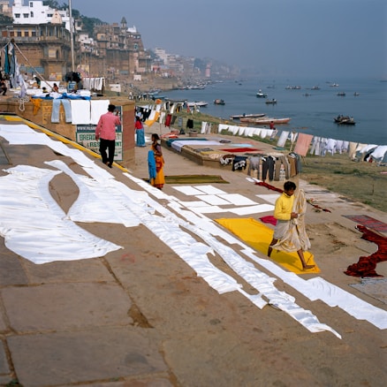 A riverside scene with people drying laundry on the ghats. White sheets are spread out on the ground, and colorful clothes hang on lines. The river has several boats floating, and the buildings line the bank, showing a dense and historic architectural style.