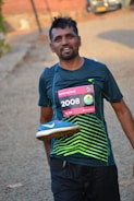 A man is smiling after completing a run, wearing a race bib numbered 2008 on his shirt. He is holding a blue running shoe with a yellow swoosh design over his shoulder. The background includes a blurred outdoor path and a vehicle in the distance.