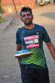 A man is smiling after completing a run, wearing a race bib numbered 2008 on his shirt. He is holding a blue running shoe with a yellow swoosh design over his shoulder. The background includes a blurred outdoor path and a vehicle in the distance.