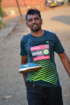 A man is smiling after completing a run, wearing a race bib numbered 2008 on his shirt. He is holding a blue running shoe with a yellow swoosh design over his shoulder. The background includes a blurred outdoor path and a vehicle in the distance.