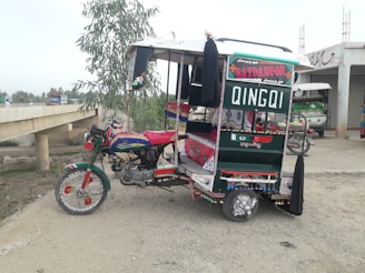 A vibrantly painted three-wheeled vehicle, known as a Qingqi rickshaw, is parked on a gravel surface. It features colorful designs and patterns with shades of red, green, blue, and white. The back displays the word 'QINGQI' and local language text. The surroundings include a tree, a partially visible bridge, and a building with rolling shutters.