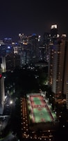A nighttime cityscape featuring tall, illuminated skyscrapers and residential buildings. In the foreground, there is a well-lit sports court. The city skyline is alive with the glow from numerous windows and colorful lights.