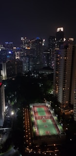 A nighttime cityscape featuring tall, illuminated skyscrapers and residential buildings. In the foreground, there is a well-lit sports court. The city skyline is alive with the glow from numerous windows and colorful lights.