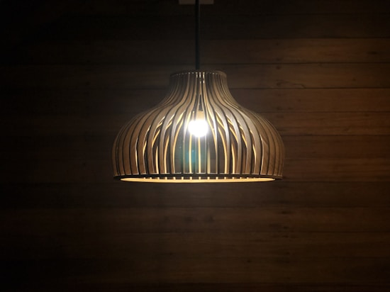 A pendant lamp with a wooden, slatted design is hanging in a dimly lit room. The light bulb inside casts a warm glow, highlighting the curved wooden slats. The background features a wooden wall with visible horizontal planks.