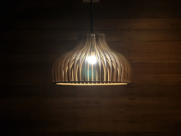 A pendant lamp with a wooden, slatted design is hanging in a dimly lit room. The light bulb inside casts a warm glow, highlighting the curved wooden slats. The background features a wooden wall with visible horizontal planks.