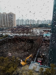 Raindrops cover a window, blurring the view of a construction site with a yellow excavator and surrounding high-rise buildings.