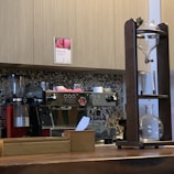 A coffee shop counter with a large espresso machine and a siphon coffee maker. The espresso machine is stainless steel with red accents and several knobs and dials. A wooden structure on the right holds glass vessels for brewing coffee. The background features light-colored cabinetry and a backsplash of small, round tiles in neutral tones.